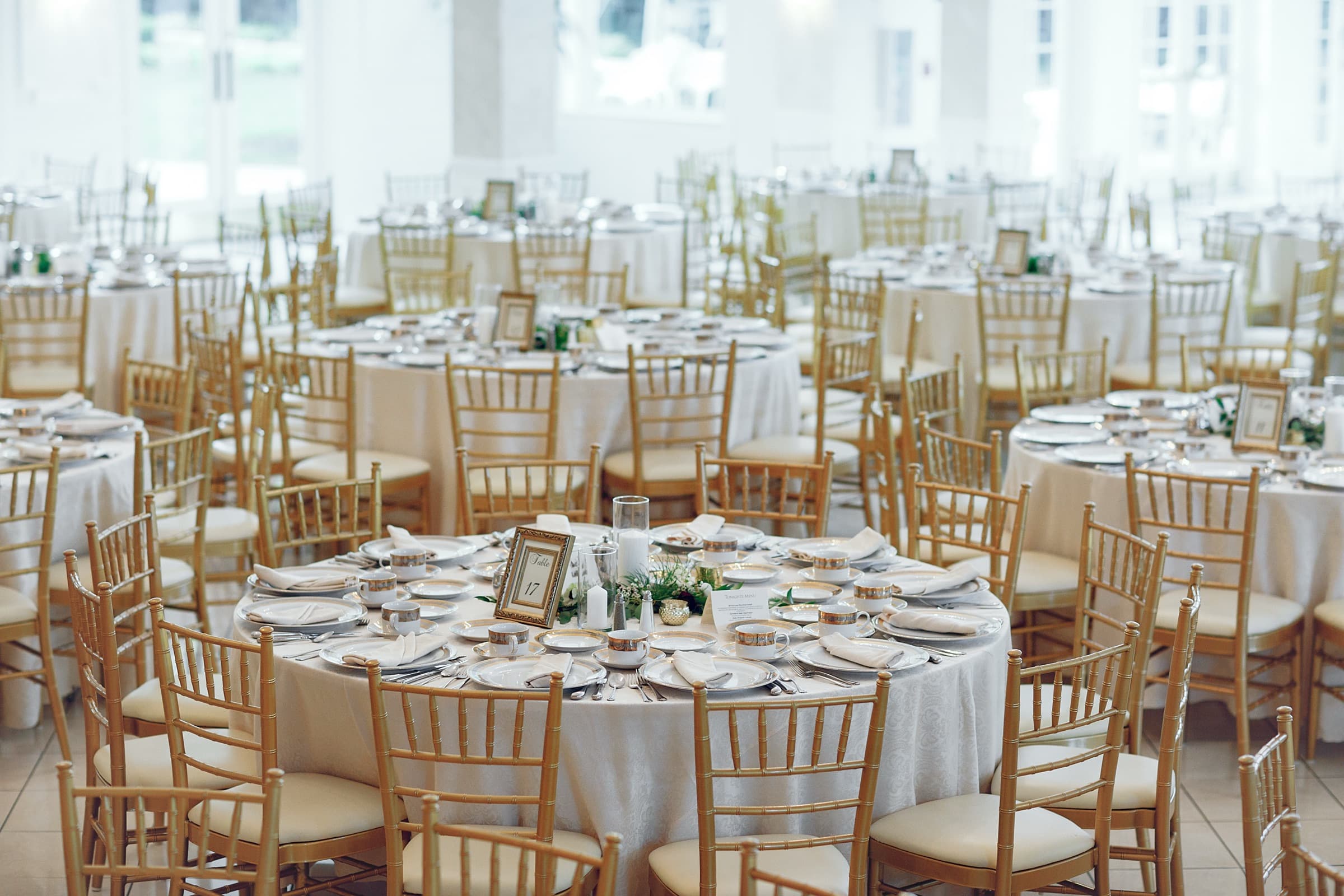 A reception hall set with round tables, white linen and gold chairs
